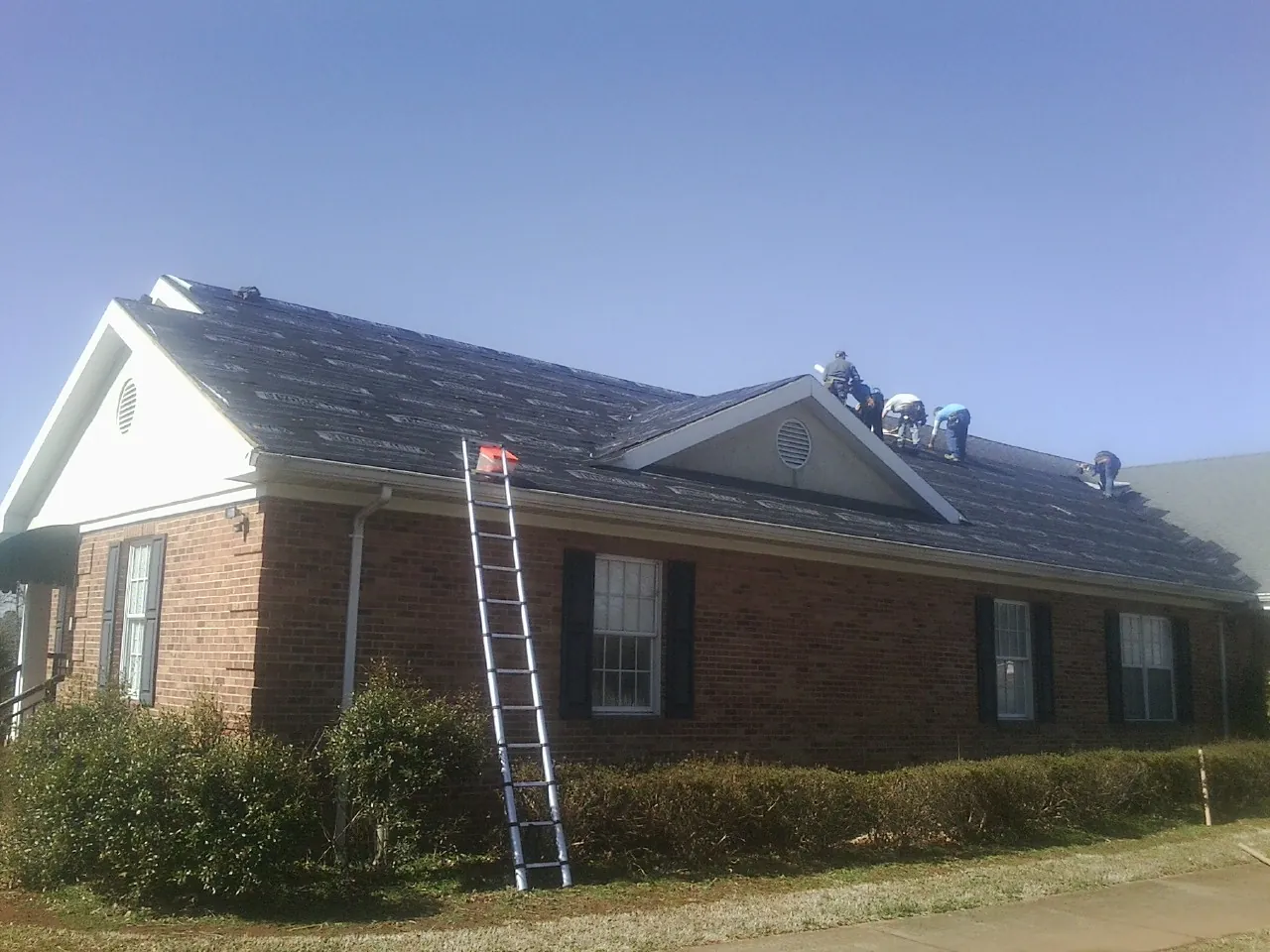 Crew tearing shingles off a brick ranch roof with a ladder set — Metal Roof Repair work by our crew in Lemoyne