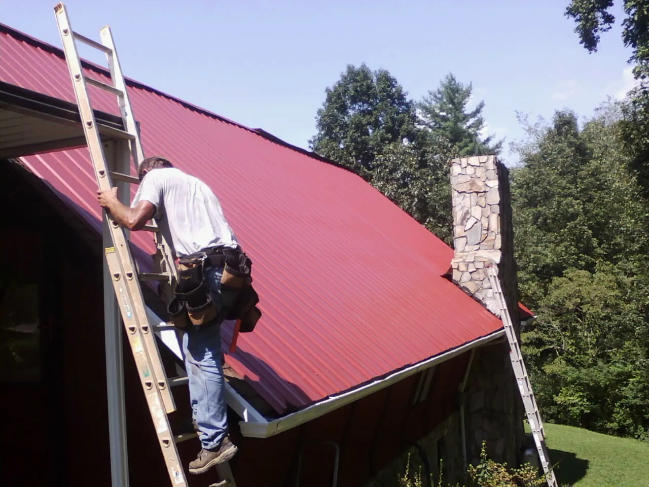 Roofer on a ladder beside a red metal roof and stone chimney — Soffit & Fascia Repair by Roofing Brothers in Lemoyne